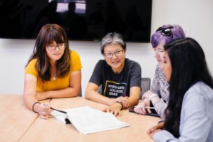 Three women students sit at a desk alongside a professor wearing a shirt that says "First Generation College Grad"