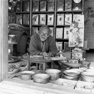 An Okinawan man in a seed store looks out at the camera, surrounded by bowls and bags of seeds. 1952.