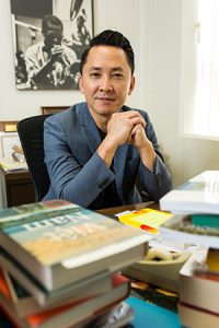 Viet Thanh Nguyen is shown seated at a desk piled with books.