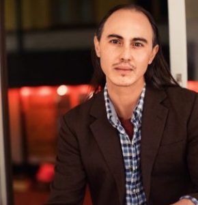 Headshot of Julian Talamantez Borlaski wearing a chequered shirt and blazer.