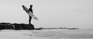 Male surfer in wet suit stands on rock with surfboard gazing into ocean