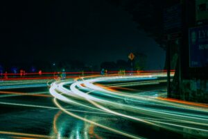 Long-exposure nighttime view of a road intersection with colorful vehicle light trails, trees, and roadside signs.